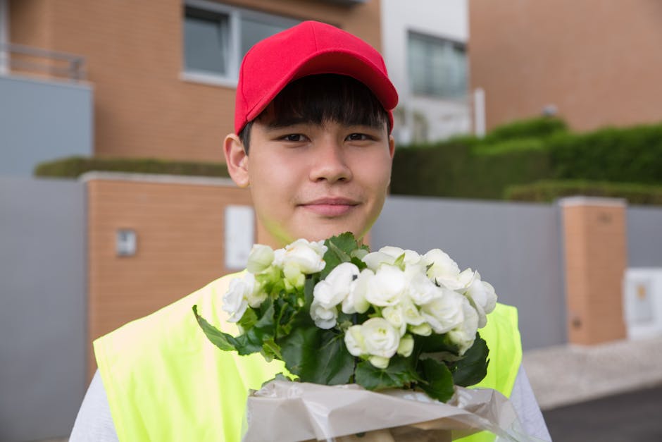 A young delivery person wearing a red cap and a high-visibility yellow vest holds a bouquet of fresh, white flowers wrapped in white paper, standing outdoors in front of modern residential buildings w