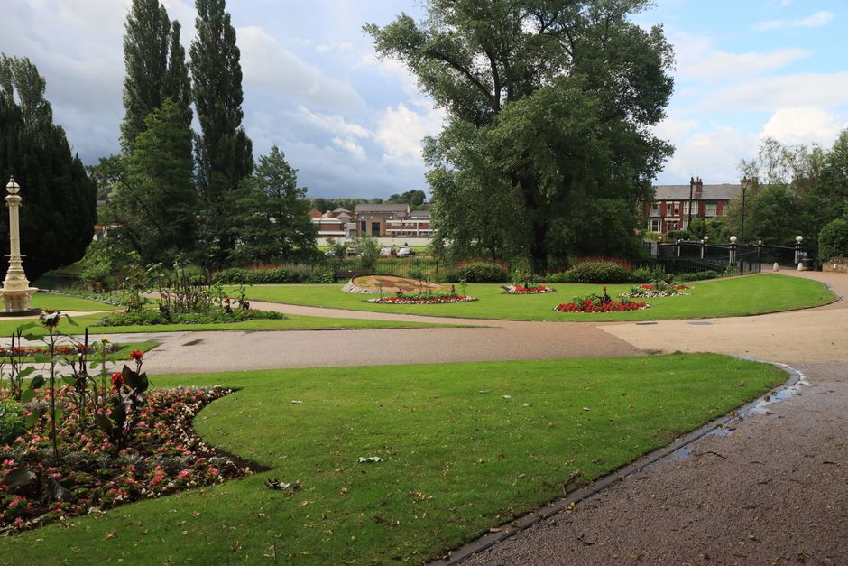 A wide view of Leyton Jubilee Park featuring manicured lawns, flowerbeds with vibrant red and pink blooms, and a large central tree with lush green foliage. The park is bordered by a curved gravel pat