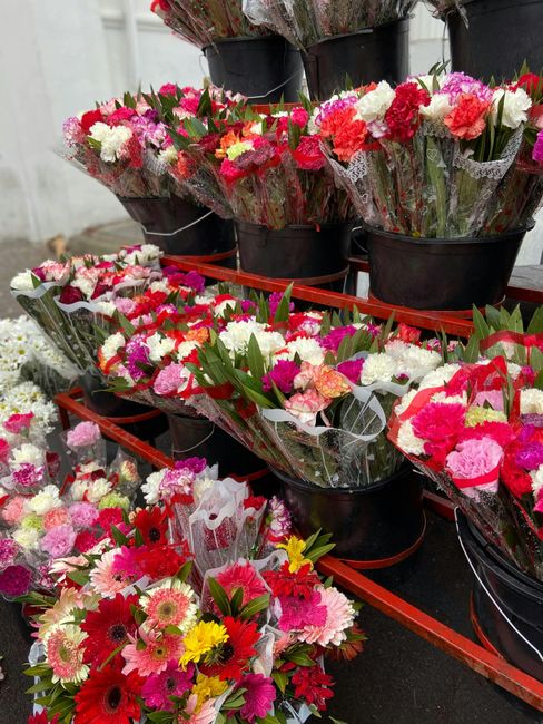 An assortment of fresh flower bouquets displayed on black plastic pots and a red trolley, featuring vibrant carnations, daisies, and roses in shades of pink, red, white, and yellow. The flowers are wr